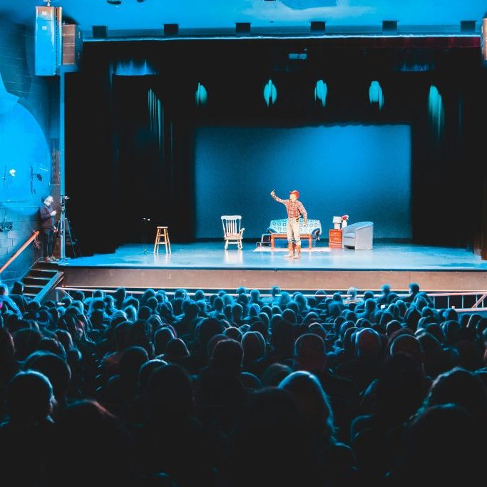 theatre packed with people watching a man perform on stage.