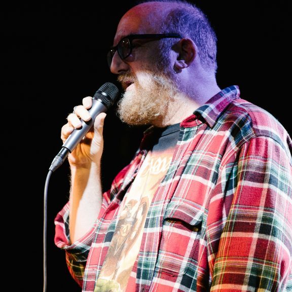 Stand up comedian on stage in a checked shirt holding a microphone